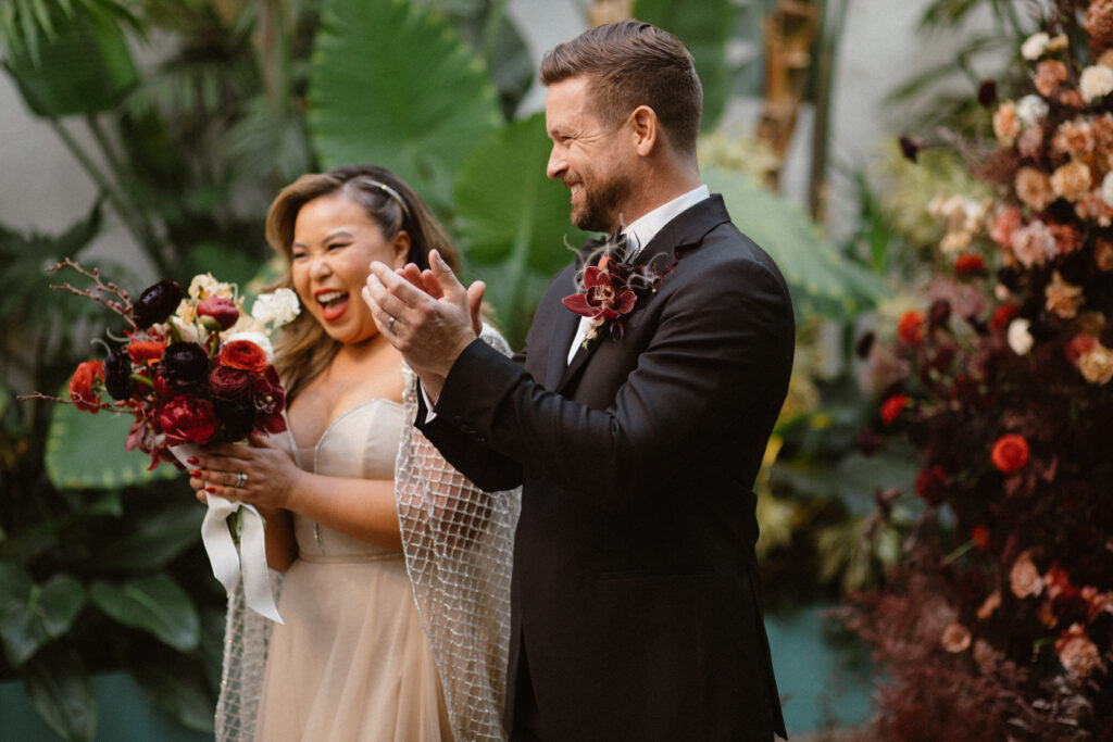a couple standing at the altar looking at the crowd celebrating and clapping because they just got married at valentine los angeles. they are surrounded by various red and rose toned florals